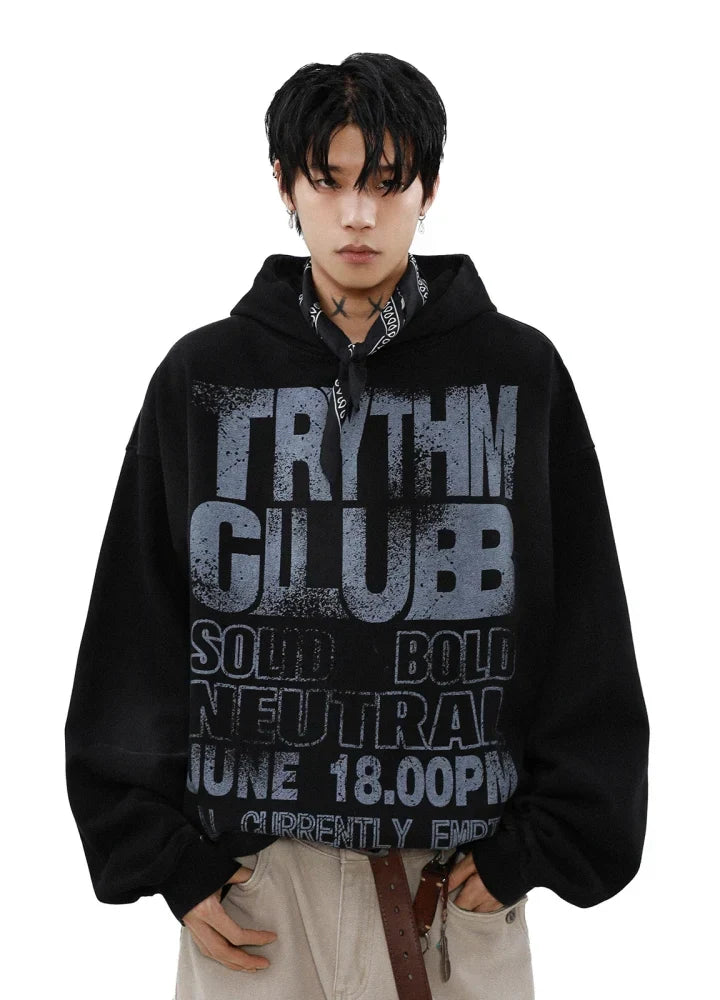 ある人物が、太字の「TRYTHM CLUBB」と「SOLID BOLD NEUTRAL」の文字が入った黒いパーカー「CANVAS HOODIE」を着ています。首には黒いバンダナを巻き、白い背景に明るい色のパンツと茶色のベルトを着用しています。.