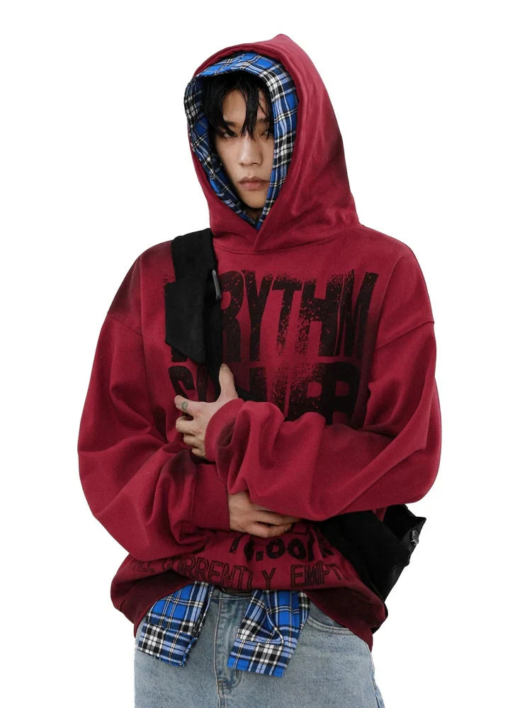 赤い文字で黒い文字が入ったフード付きのキャンバス パーカーを着て、青いチェックのシャツを重ね着し、ライトブルーのジーンズをはき、手を組んで下を向いて立っている人物。.